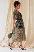 A woman wearing black sandals and a sheer, knee-length dress with gold floral embroidery over a black slip holds the Sarvin Golden & Black Italian Lace Pouch Bag, standing before a beige fabric backdrop.