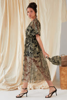 A woman in a sheer embroidered dress with black heels stands sideways, holding the Sarvin Golden & Black Italian Lace Pouch Bag. Beige draped fabric and a woven basket are seen in the background.