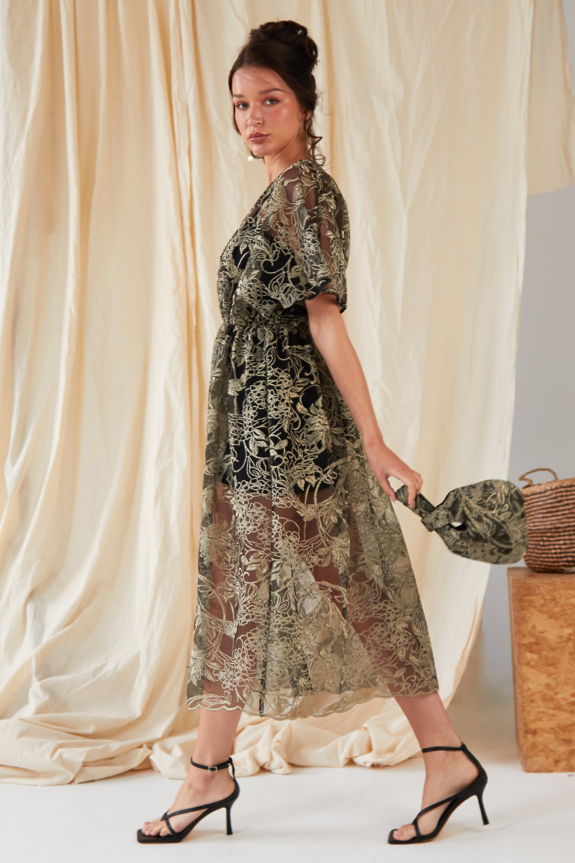A woman in a sheer embroidered dress with black heels stands sideways, holding the Sarvin Golden & Black Italian Lace Pouch Bag. Beige draped fabric and a woven basket are seen in the background.