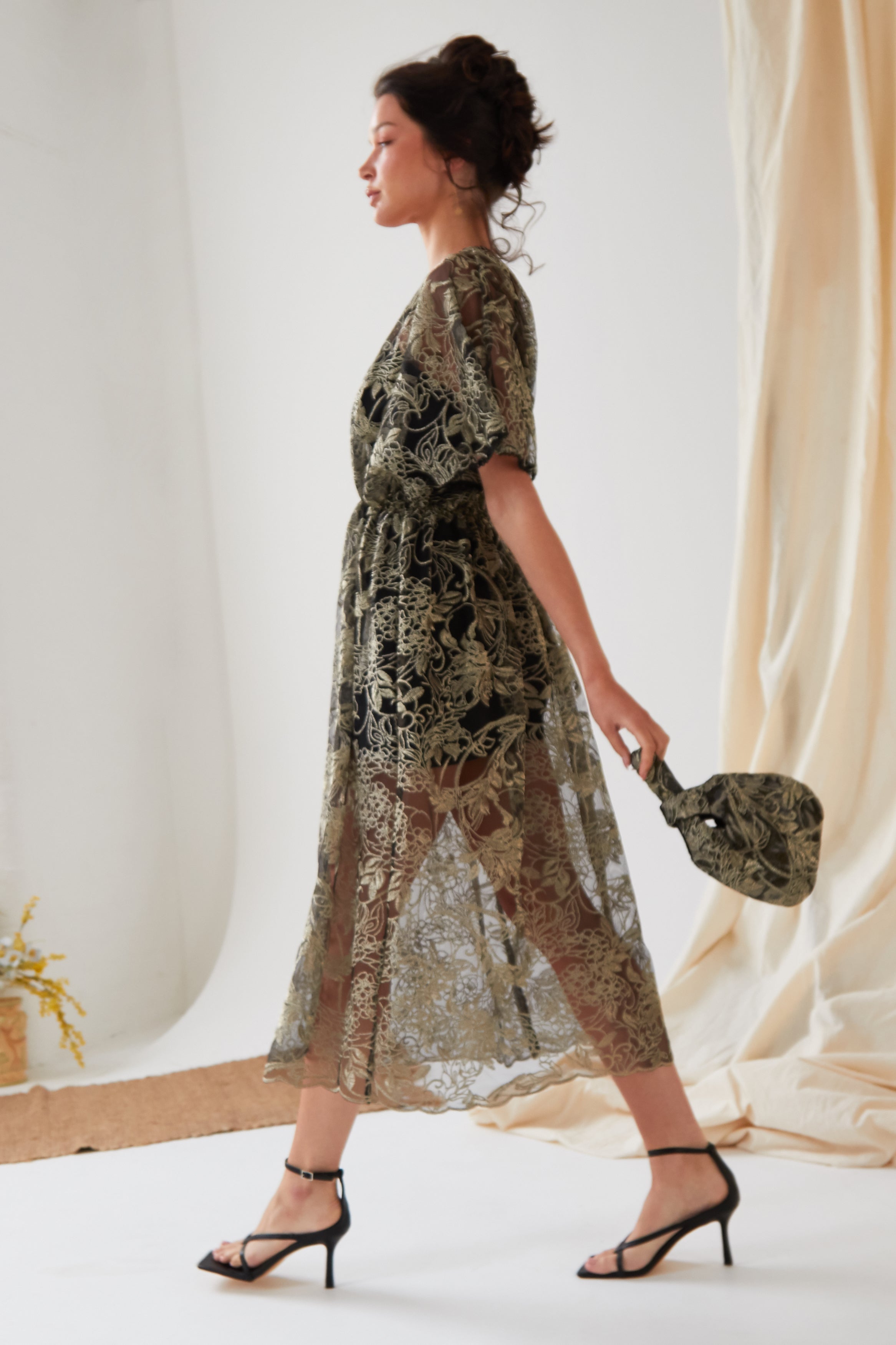 A woman in profile walks indoors, wearing a sheer patterned dress over a slip and black strappy heels. She holds the Sarvin Golden & Black Italian Lace Pouch Bag. Light drapes and a small plant appear in the background.