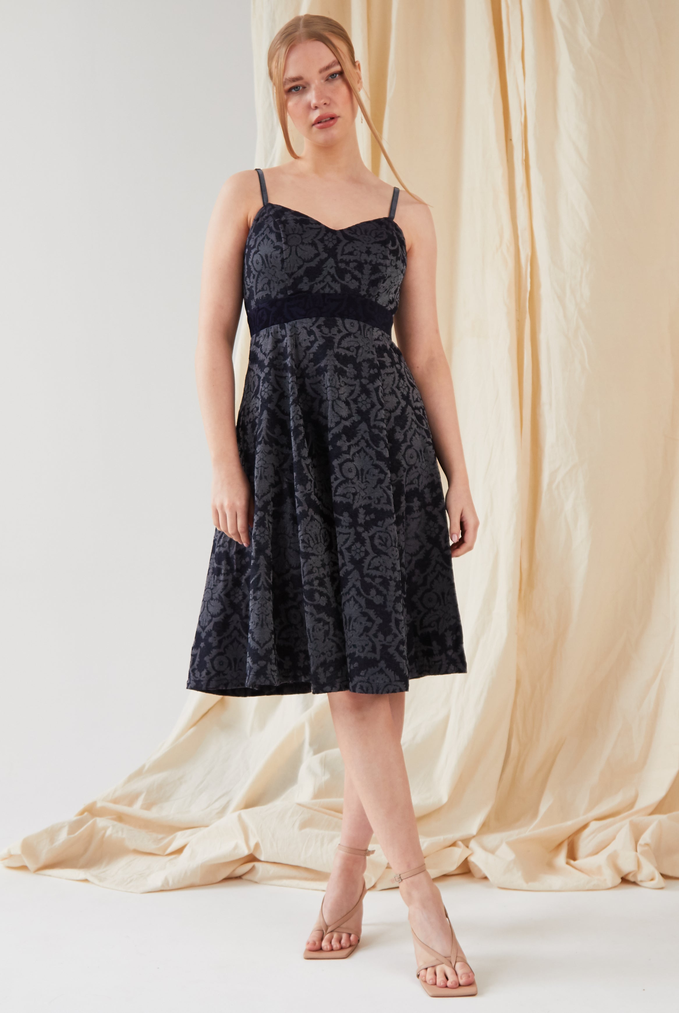 A person stands before a beige draped backdrop, wearing Sarvin's sleeveless, knee-length Jacquard Skater Dress with a floral pattern and nude heeled sandals.