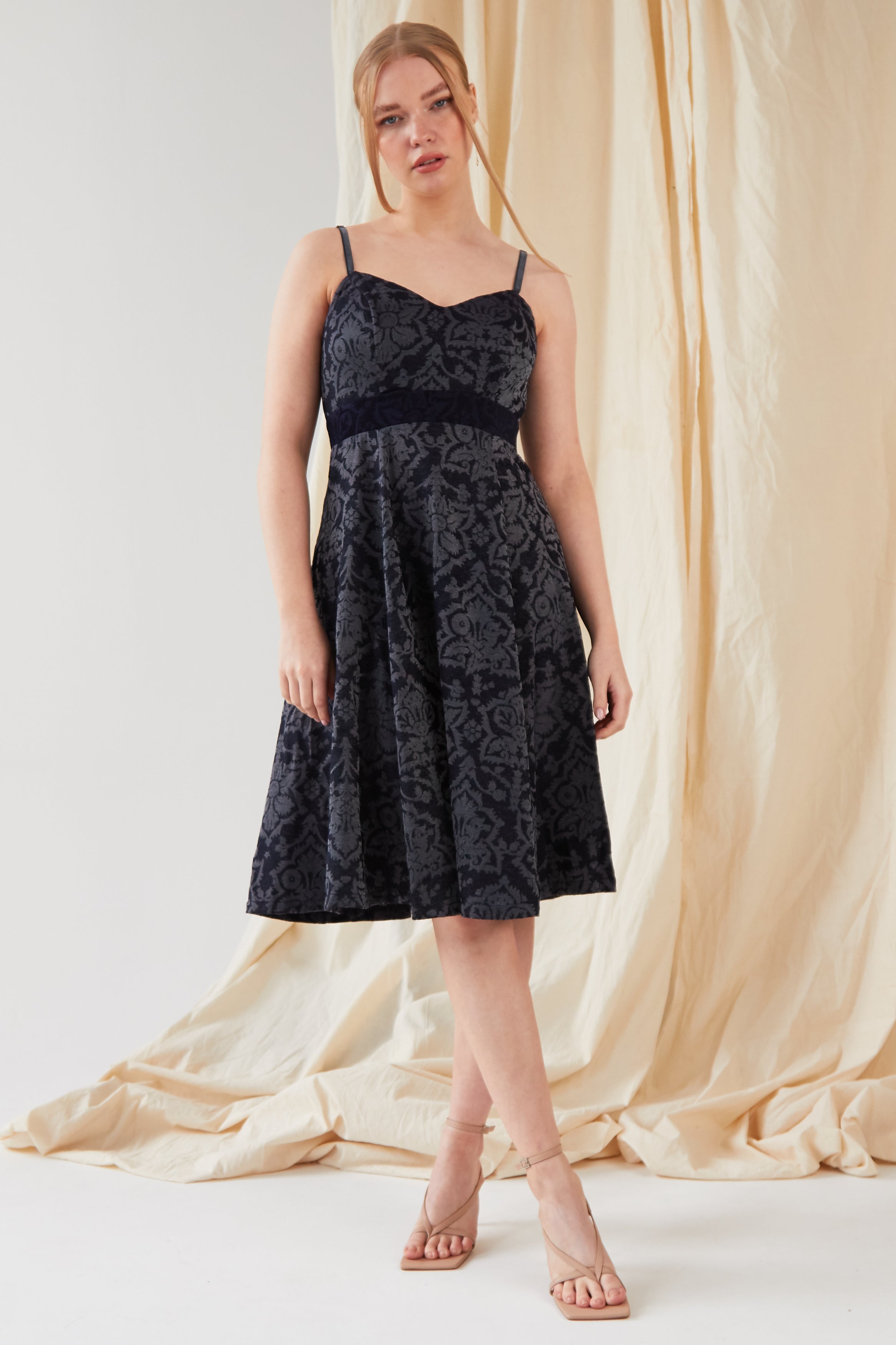 A person stands before a beige draped backdrop, wearing Sarvin's sleeveless, knee-length Jacquard Skater Dress with a floral pattern and nude heeled sandals.