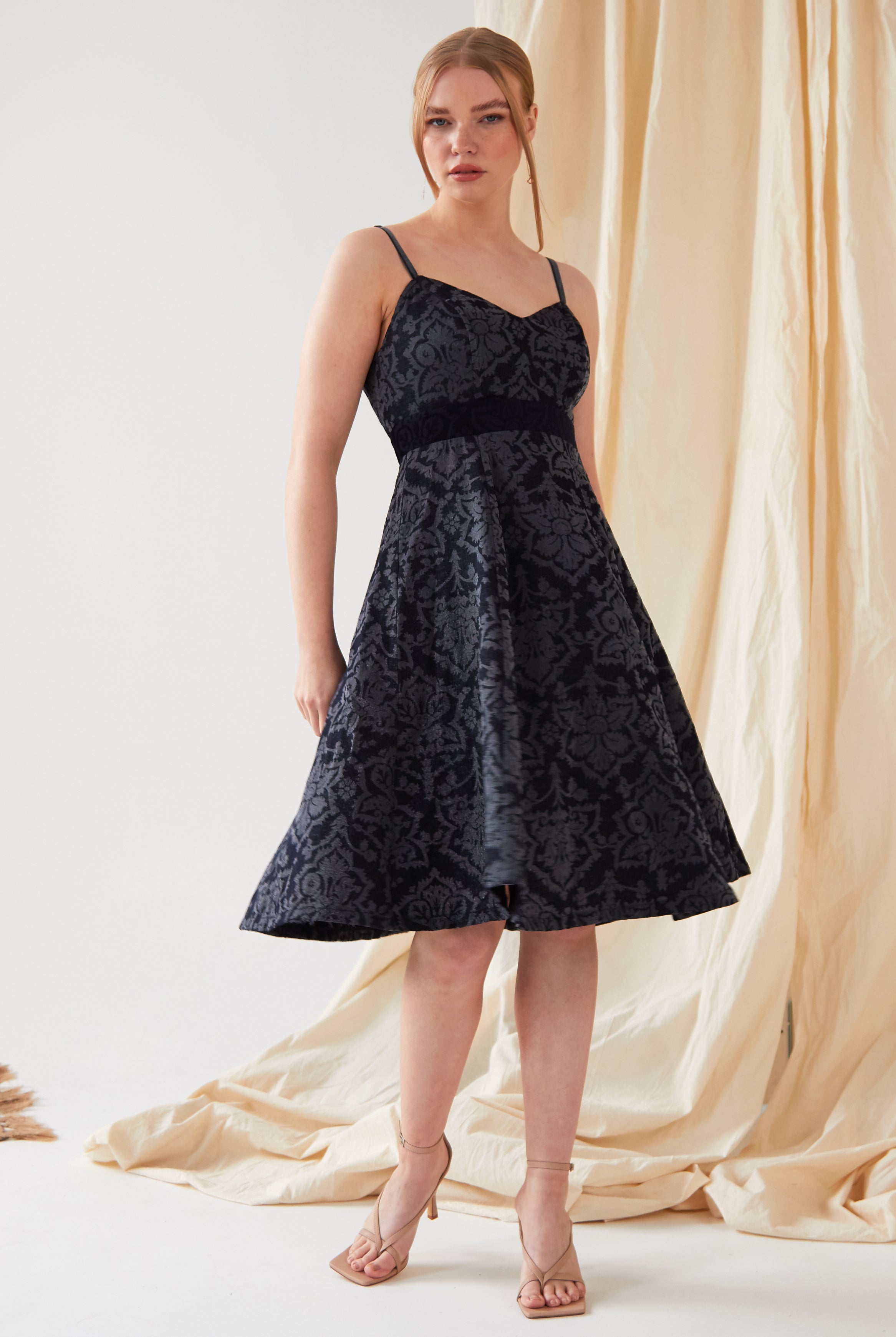 A woman with her hair tied back stands before a beige curtain, wearing the Sarvin Jacquard Skater Dress—navy, sleeveless, knee-length with thin velvet straps and a textured floral pattern—paired with nude high-heeled sandals.
