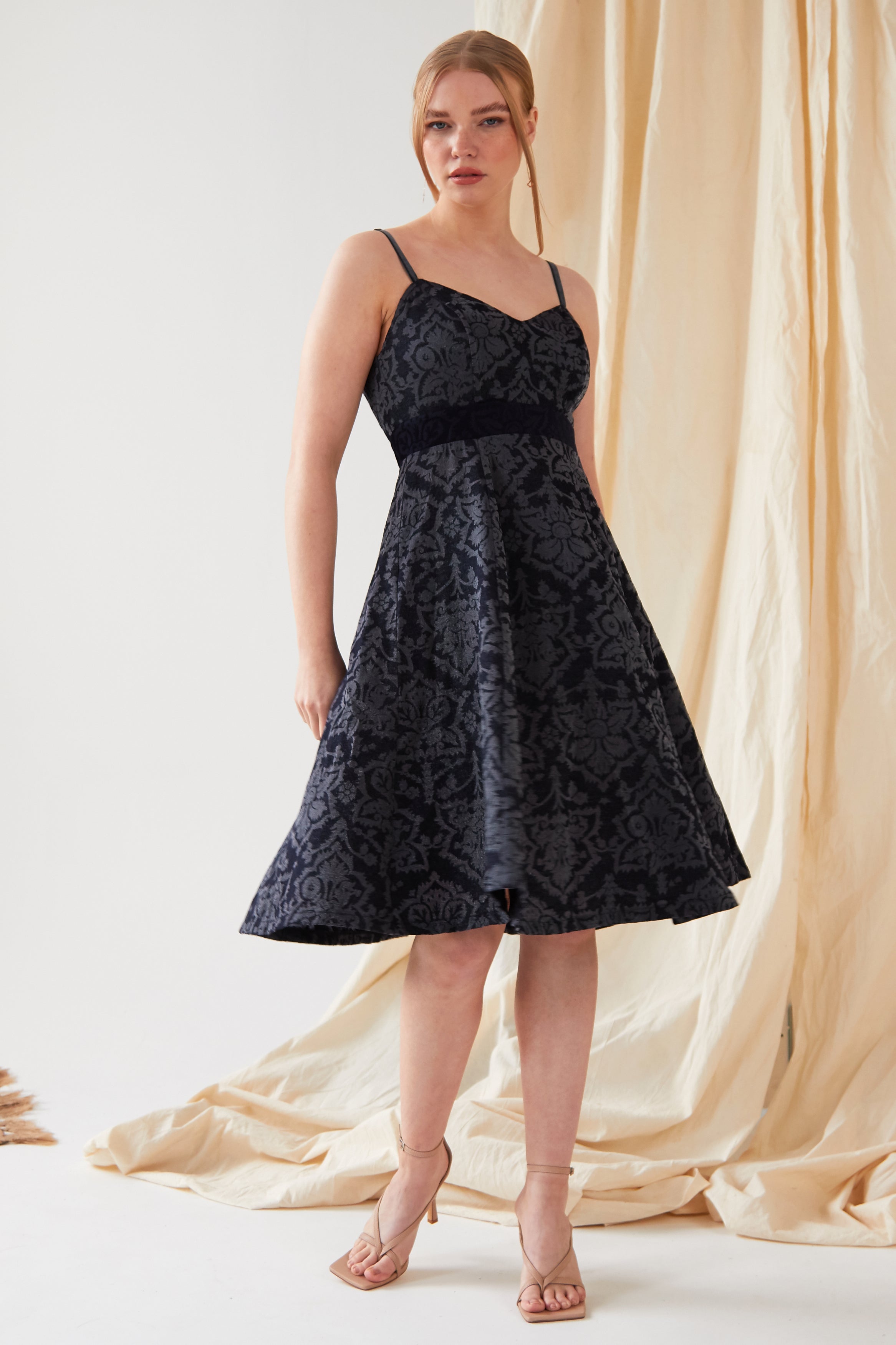 A woman with her hair tied back stands before a beige curtain, wearing the Sarvin Jacquard Skater Dress—navy, sleeveless, knee-length with thin velvet straps and a textured floral pattern—paired with nude high-heeled sandals.