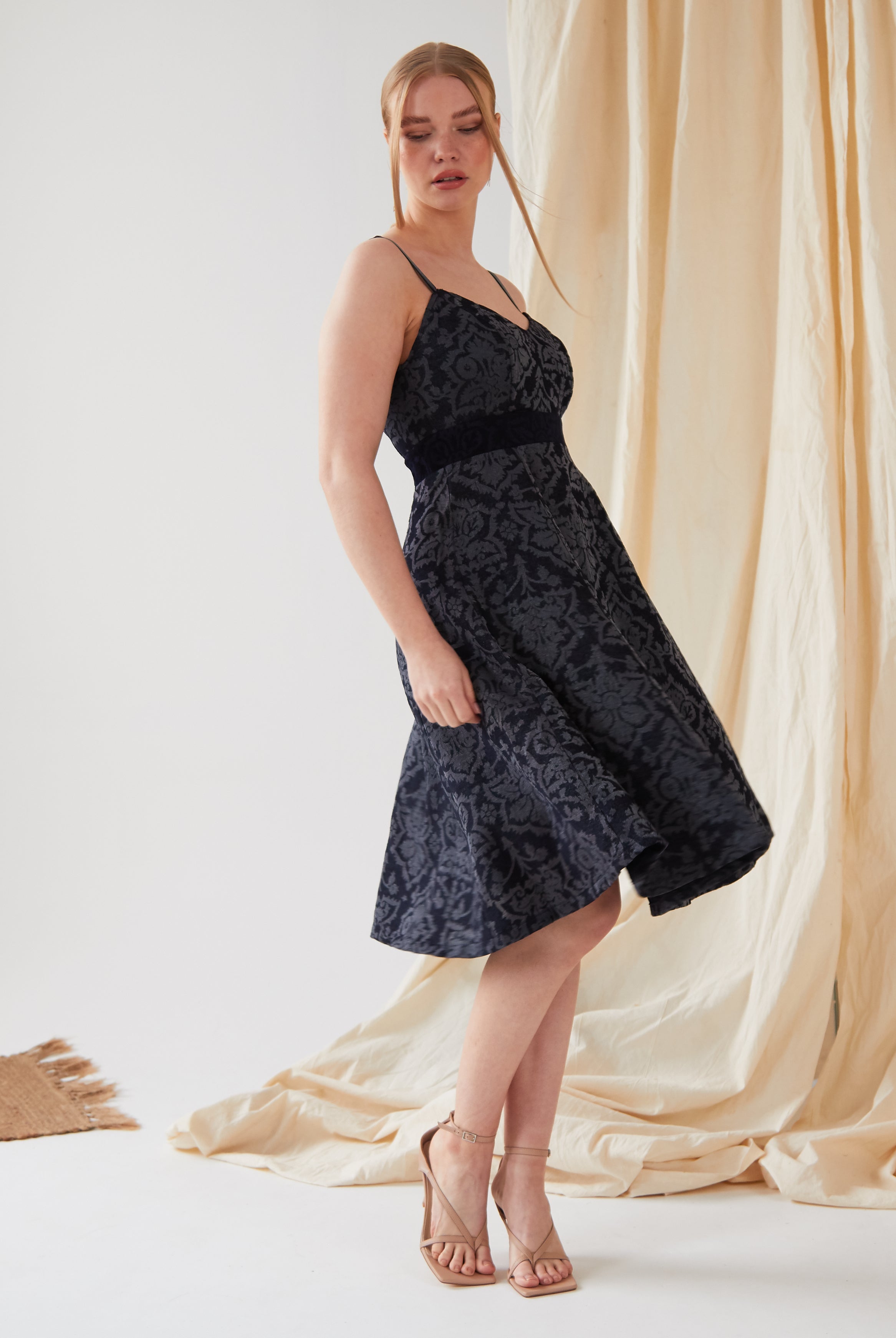A woman stands indoors wearing the Sarvin Jacquard Skater Dress—sleeveless, knee-length, with a textured floral pattern and velvet straps—paired with strappy nude heels. Light fabric drapes behind her, and part of a woven rug is visible on the floor.