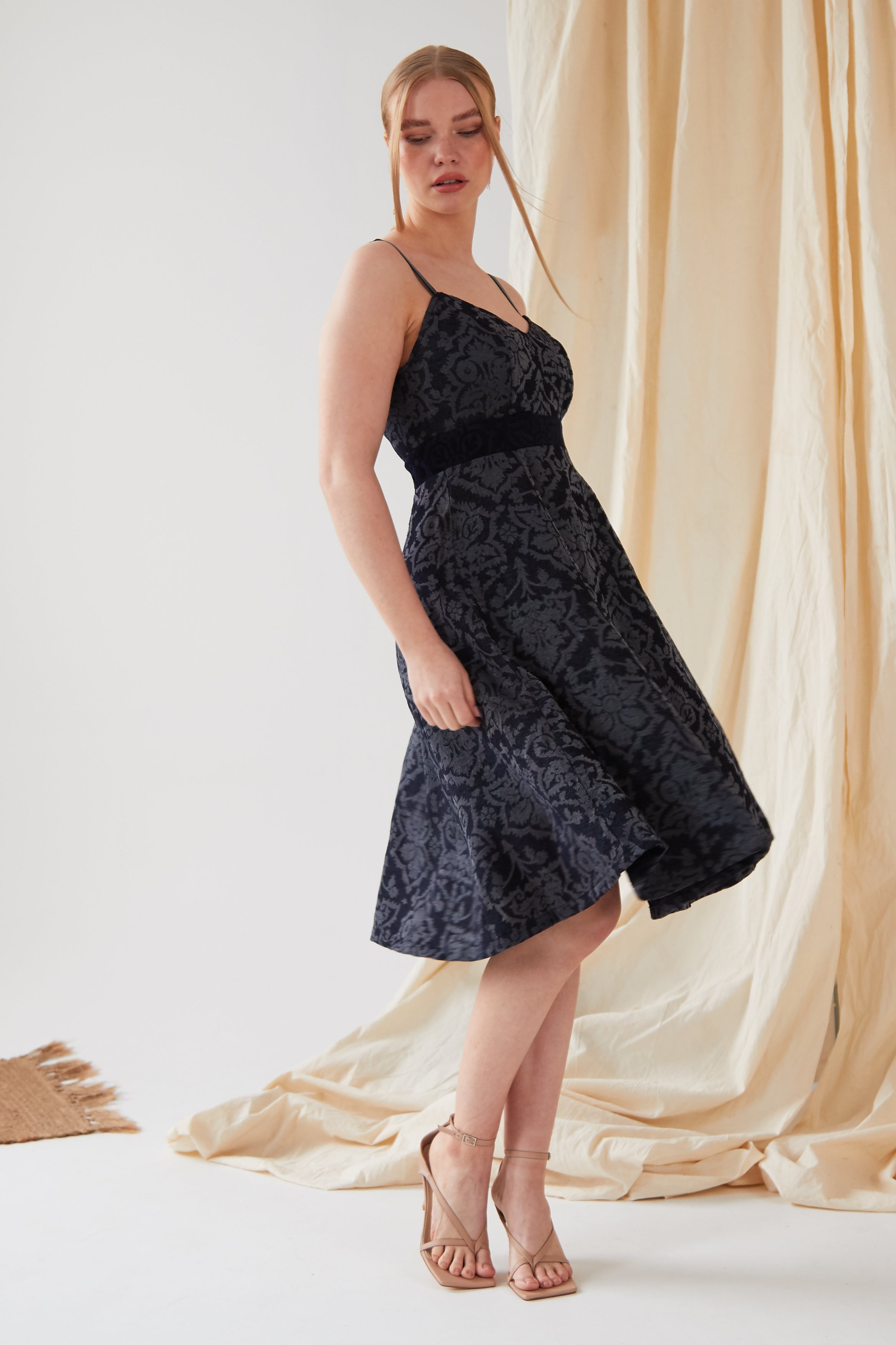 A woman stands indoors wearing the Sarvin Jacquard Skater Dress—sleeveless, knee-length, with a textured floral pattern and velvet straps—paired with strappy nude heels. Light fabric drapes behind her, and part of a woven rug is visible on the floor.