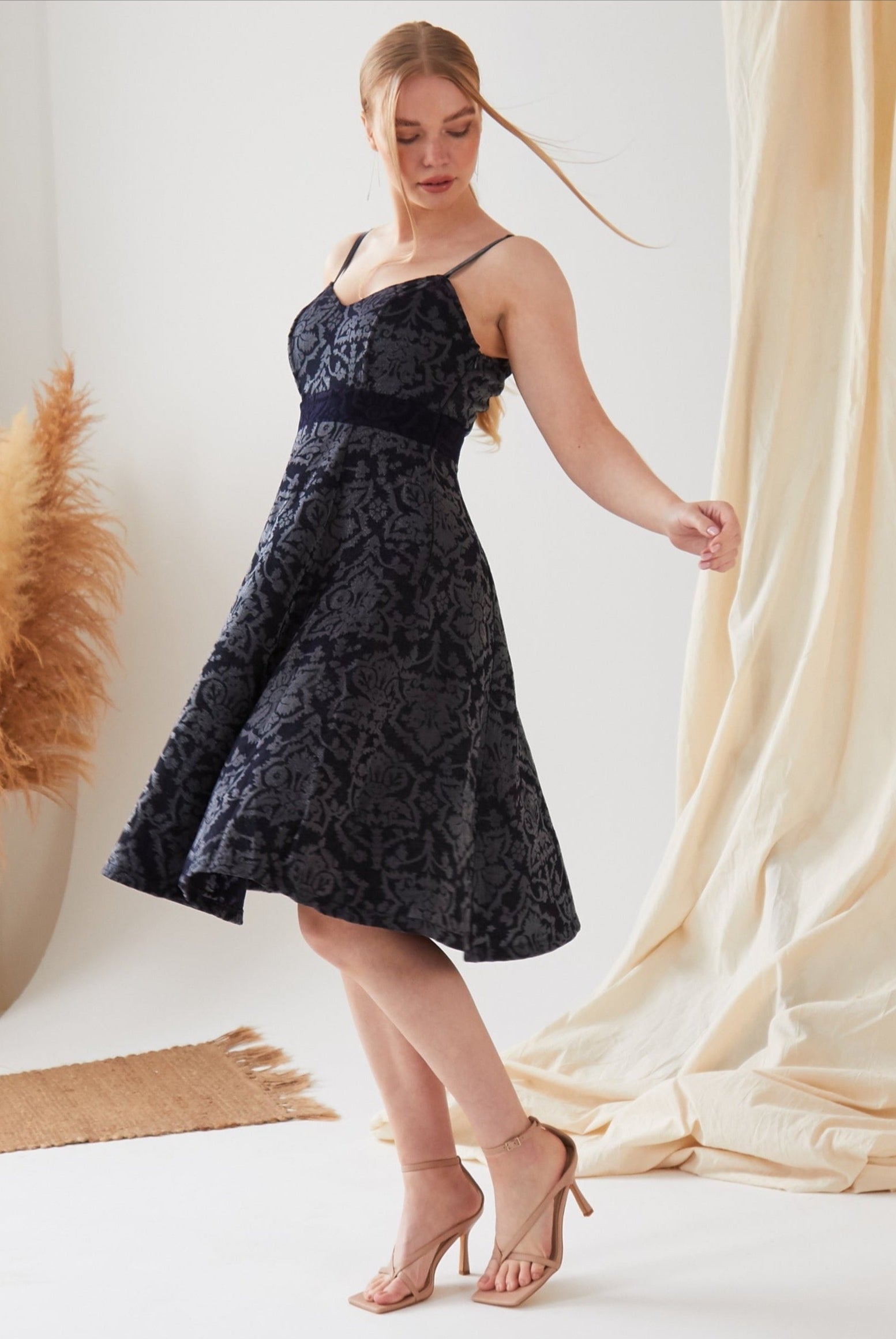 A woman wears the Sarvin Jacquard Skater Dress, a black, sleeveless, eco-friendly dress with a floral pattern. She stands on one foot in a light room featuring beige curtains and pampas grass decor.