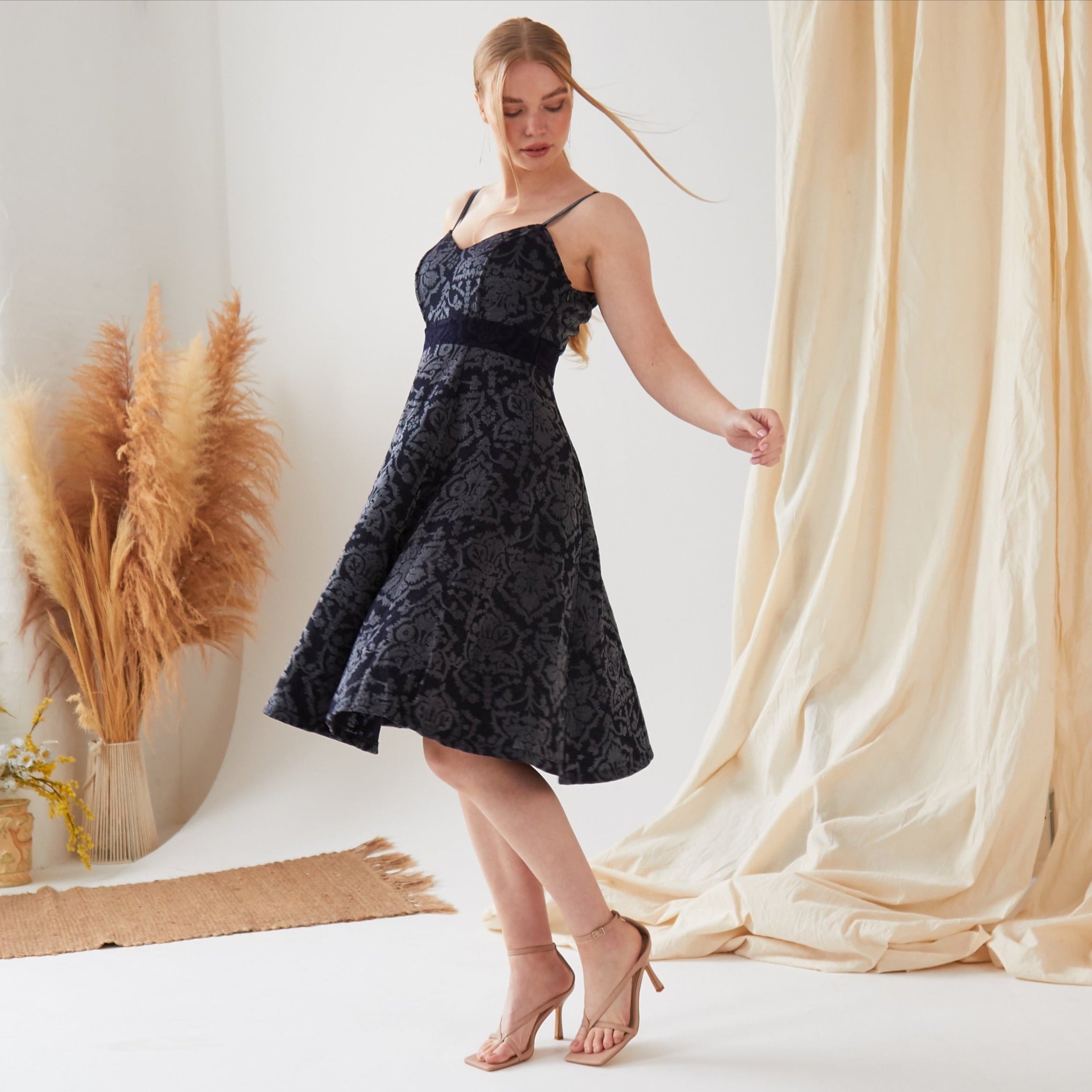 A woman wears the Sarvin Jacquard Skater Dress, a black, sleeveless, eco-friendly dress with a floral pattern. She stands on one foot in a light room featuring beige curtains and pampas grass decor.