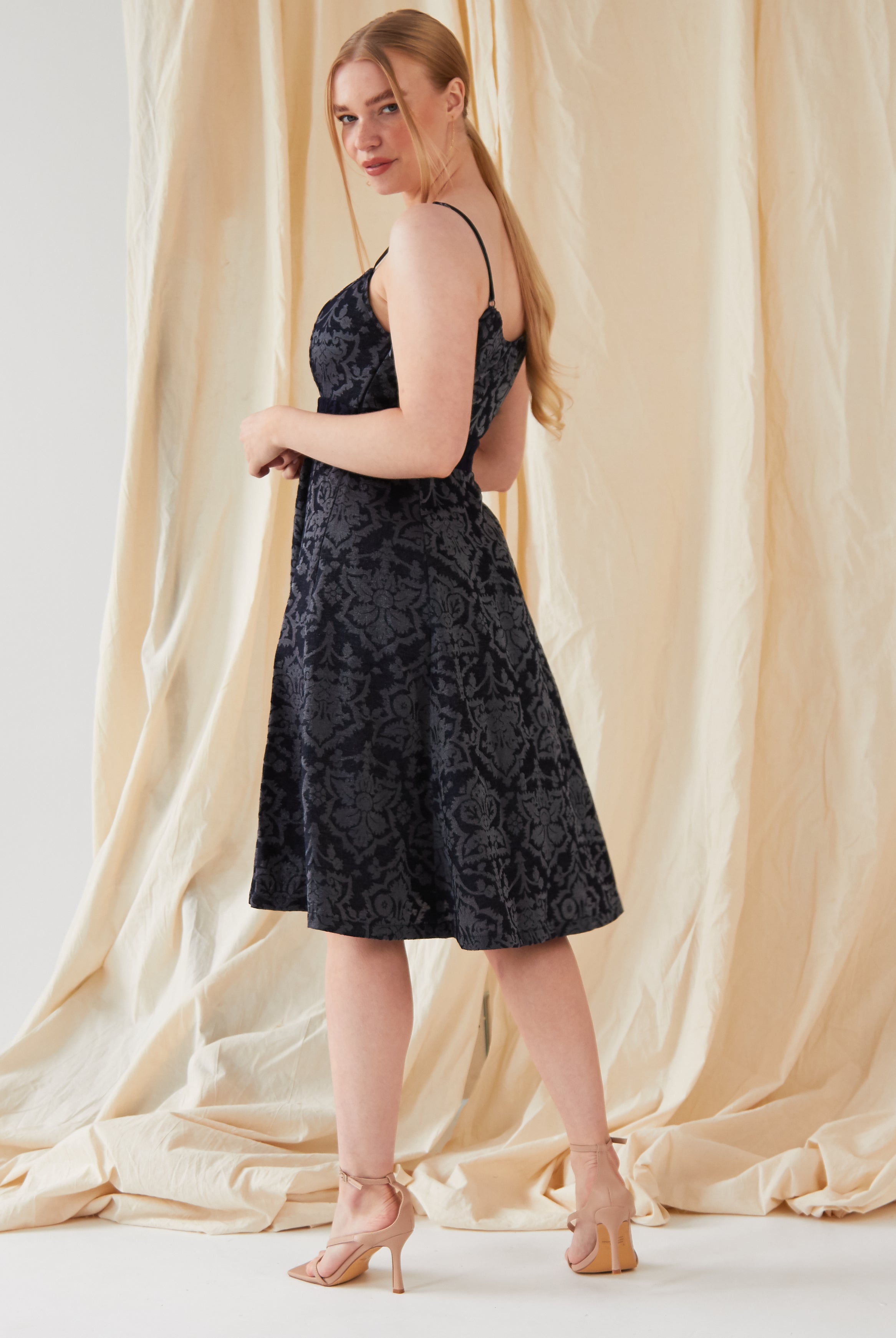 A woman with long blonde hair stands before a cream draped backdrop, wearing the Sarvin Jacquard Skater Dress with a floral pattern and nude high-heeled sandals, looking over her shoulder.