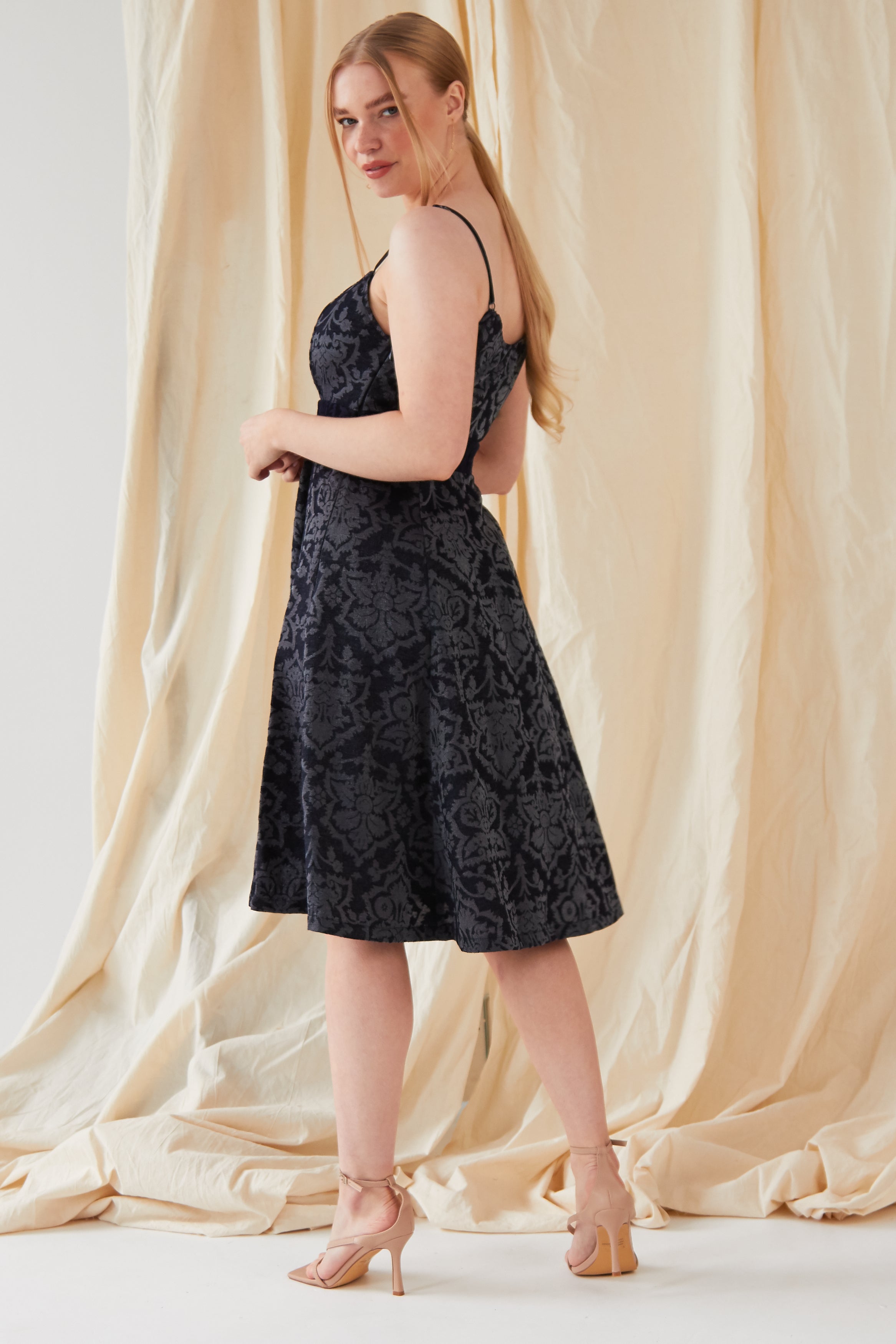 A woman with long blonde hair stands before a cream draped backdrop, wearing the Sarvin Jacquard Skater Dress with a floral pattern and nude high-heeled sandals, looking over her shoulder.