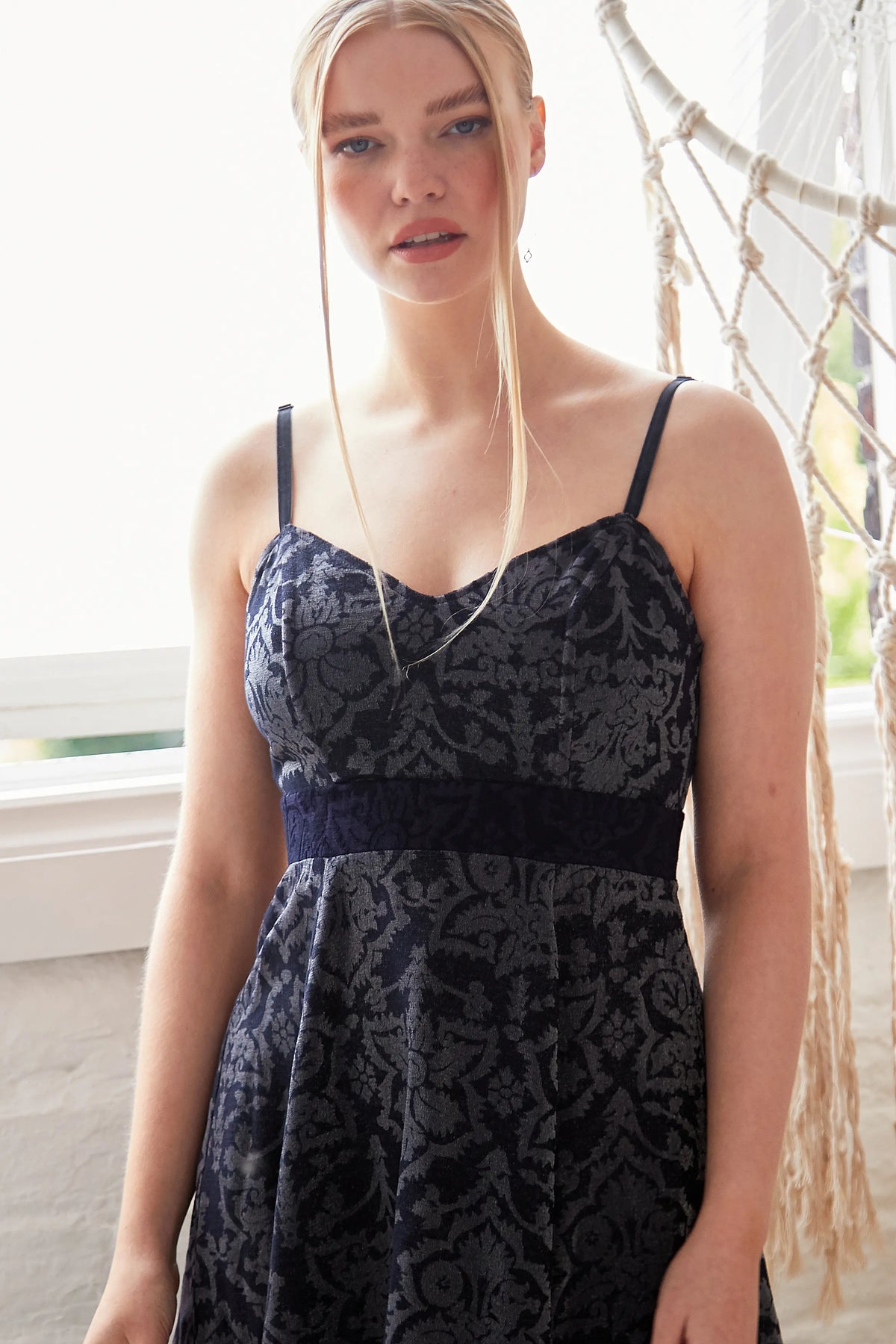 A woman with long blonde hair wears the Sarvin Jacquard Skater Dress, featuring thin velvet straps. She stands indoors by a window, surrounded by white walls and macramé decor in the background.