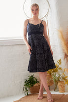 A woman stands indoors in front of a white brick wall, surrounded by plants and dried grasses, wearing the Sarvin Jacquard Skater Dress with velvet straps and strappy heels.