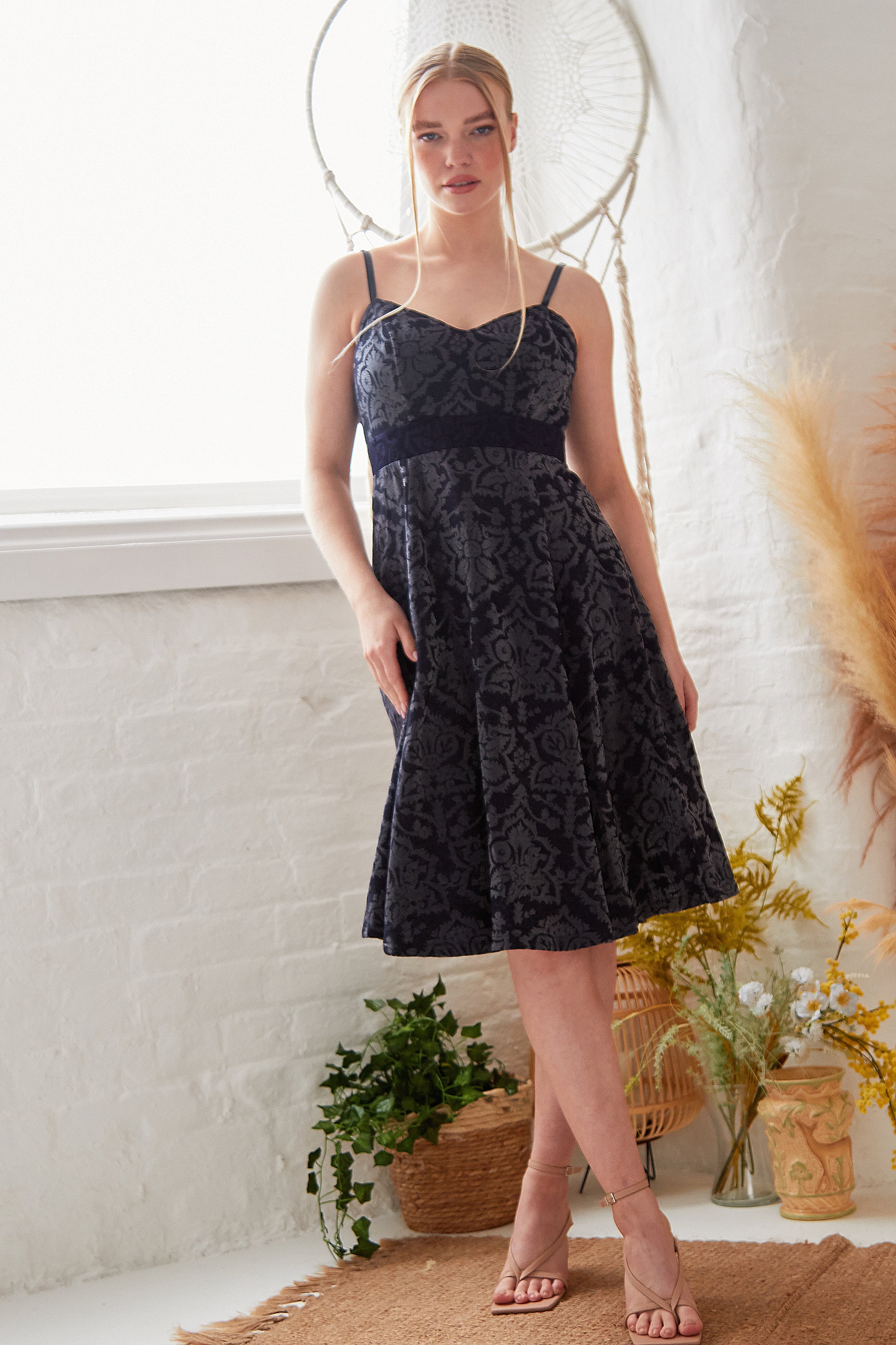 A woman stands indoors in front of a white brick wall, surrounded by plants and dried grasses, wearing the Sarvin Jacquard Skater Dress with velvet straps and strappy heels.