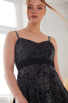 A woman with fair skin and strawberry blonde hair wears the Sarvin Jacquard Skater Dress with velvet straps. She stands indoors in natural light, gazing slightly down at the camera.