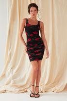 A woman stands before cream draped fabric, wearing the Sarvin Ruched Bodycon Mini Dress—a UK-made, sleeveless black mini dress with red rose floral print—and black strappy heels. She poses with one hand on her hip, looking at the camera.