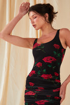 A woman poses in front of a cream curtain wearing the Sarvin Ruched Bodycon Mini Dress, featuring a red rose floral print, with one arm raised and her head tilted back.