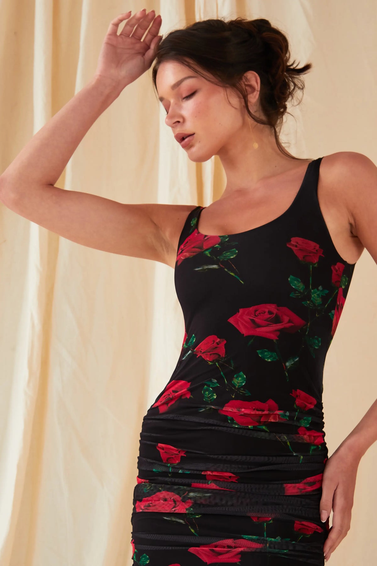 A woman poses in front of a cream curtain wearing the Sarvin Ruched Bodycon Mini Dress, featuring a red rose floral print, with one arm raised and her head tilted back.