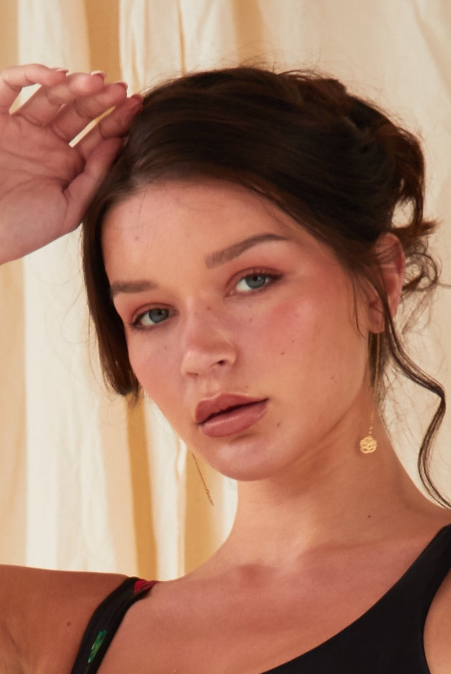 A woman with light skin and brown hair poses before a beige fabric backdrop wearing a black sleeveless top and Sarvin Flower Shape 18K Gold Drop Earrings, one hand raised near her head.