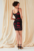 A woman faces away from the camera before beige draped fabric, wearing the Sarvin Ruched Bodycon Mini Dress—a UK-made mini dress with a red rose floral print—paired with black high-heeled sandals.