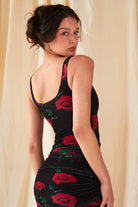 A person with dark hair in a loose updo models the Sarvin Ruched Bodycon Mini Dress, made in the UK and featuring a red rose floral print, as they stand before a light fabric backdrop and glance over their shoulder at the camera.