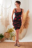 A woman stands indoors wearing the Sarvin Ruched Bodycon Mini Dress—sleeveless, black with a red rose floral print—paired with black heels. She is surrounded by boho decor like dried plants, baskets, and a woven wall hanging.