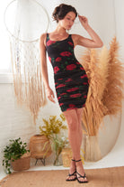 Indoors, a woman wears the Sarvin Ruched Bodycon Mini Dress, featuring a red rose floral print. She poses with one hand near her hair, amid decorative plants, a woven rug, and a large dreamcatcher, paired with black heeled sandals.