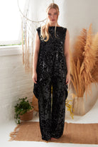 A woman stands indoors on a brown rug, wearing Sarvin's handmade in the UK Sequin Velvet Flared Trousers. Behind her are tall dried plants in vases, a white wall, and a window letting in natural light.