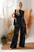 A woman stands indoors on a woven rug, wearing Sarvin's Sequin Velvet Flared Trousers. She is surrounded by neutral decor, including a large dreamcatcher, pampas grass, and houseplants in wicker baskets.