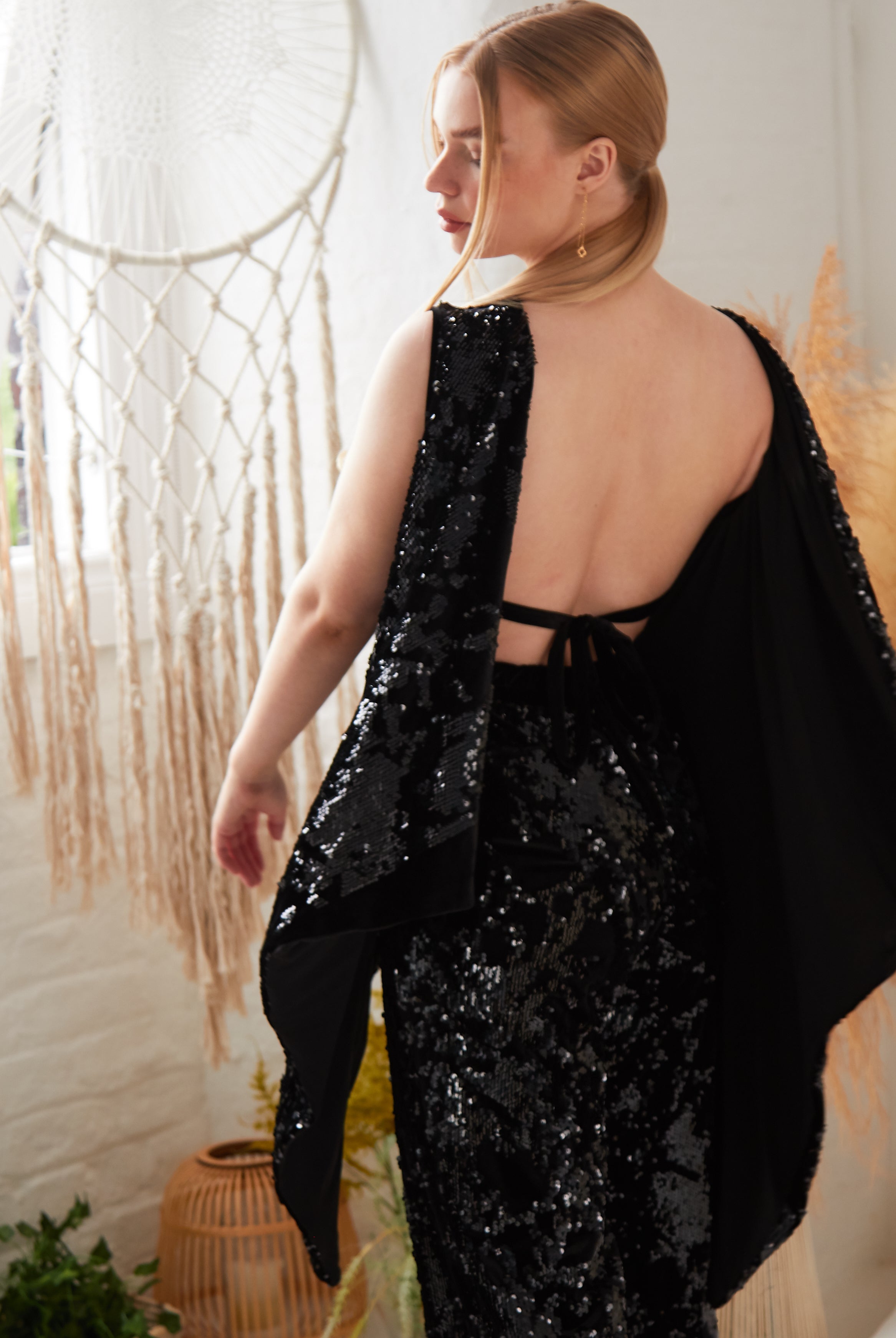 A woman with blonde hair models Sarvin's Sequin Velvet Flared Trousers indoors, her back to the camera. A macramé wall hanging and dried plants in the background evoke a chic, handcrafted UK-inspired ambiance.