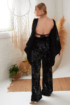 A woman stands in a bright room with her back to the camera, wearing Sarvin’s Sequin Velvet Flared Trousers and a black sequined open-back top. The decor features a dreamcatcher, pampas grass, a rug, and potted plants.