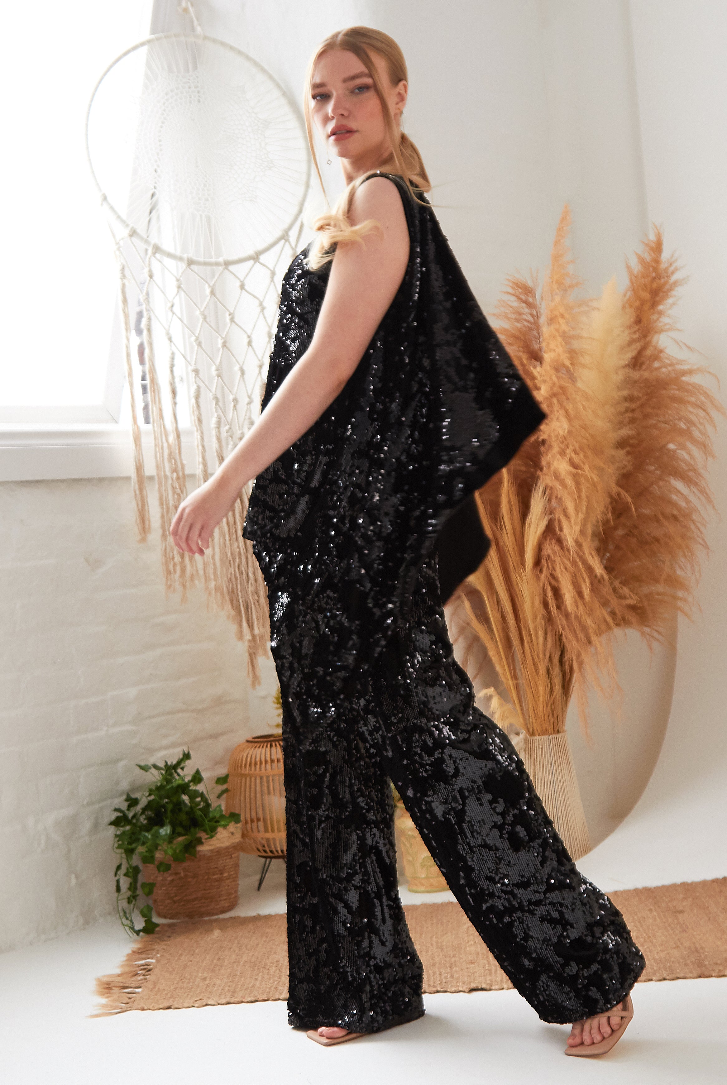 A woman indoors wears Sarvin Sequin Velvet Flared Trousers. Behind her are pampas grass, a wicker chair, a potted plant, a white brick wall, and a large dreamcatcher with light streaming through the window.