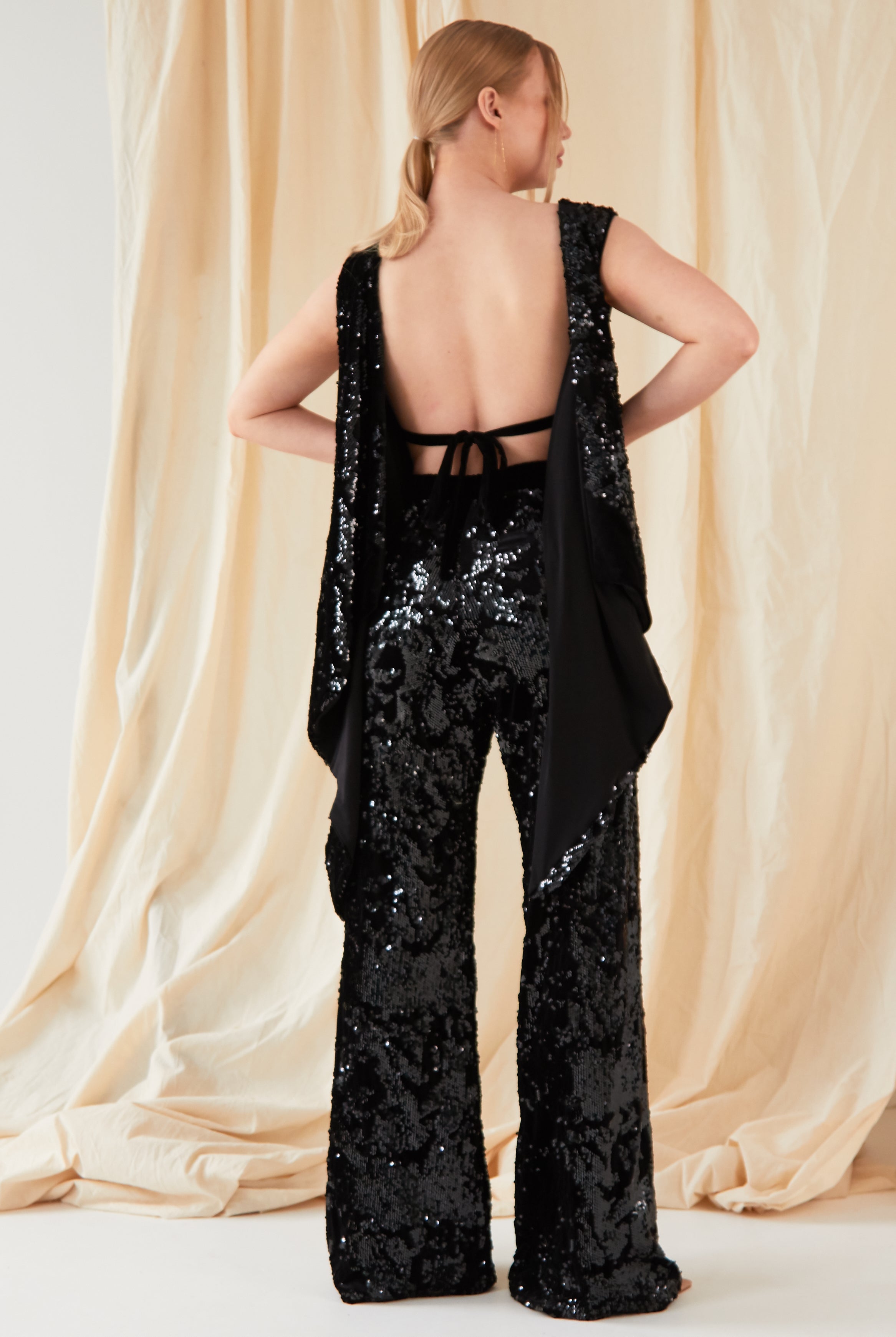 A person faces away from the camera, wearing Sarvin’s handmade Sequin Velvet Flared Trousers and a matching backless sequin top, posed before light beige draped fabric.