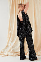 A person stands before beige draped fabric, wearing a loose black sequin top with an open back and Sarvin Sequin Velvet Flared Trousers. Their arm is raised behind their head, gaze over one shoulder.