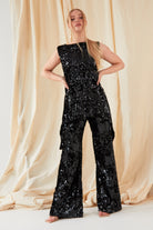 A person stands before draped beige fabric, wearing a sleeveless black sequin tunic and Sarvin’s handmade in the UK Sequin Velvet Flared Trousers. Their long blonde hair is styled in a sleek low ponytail.