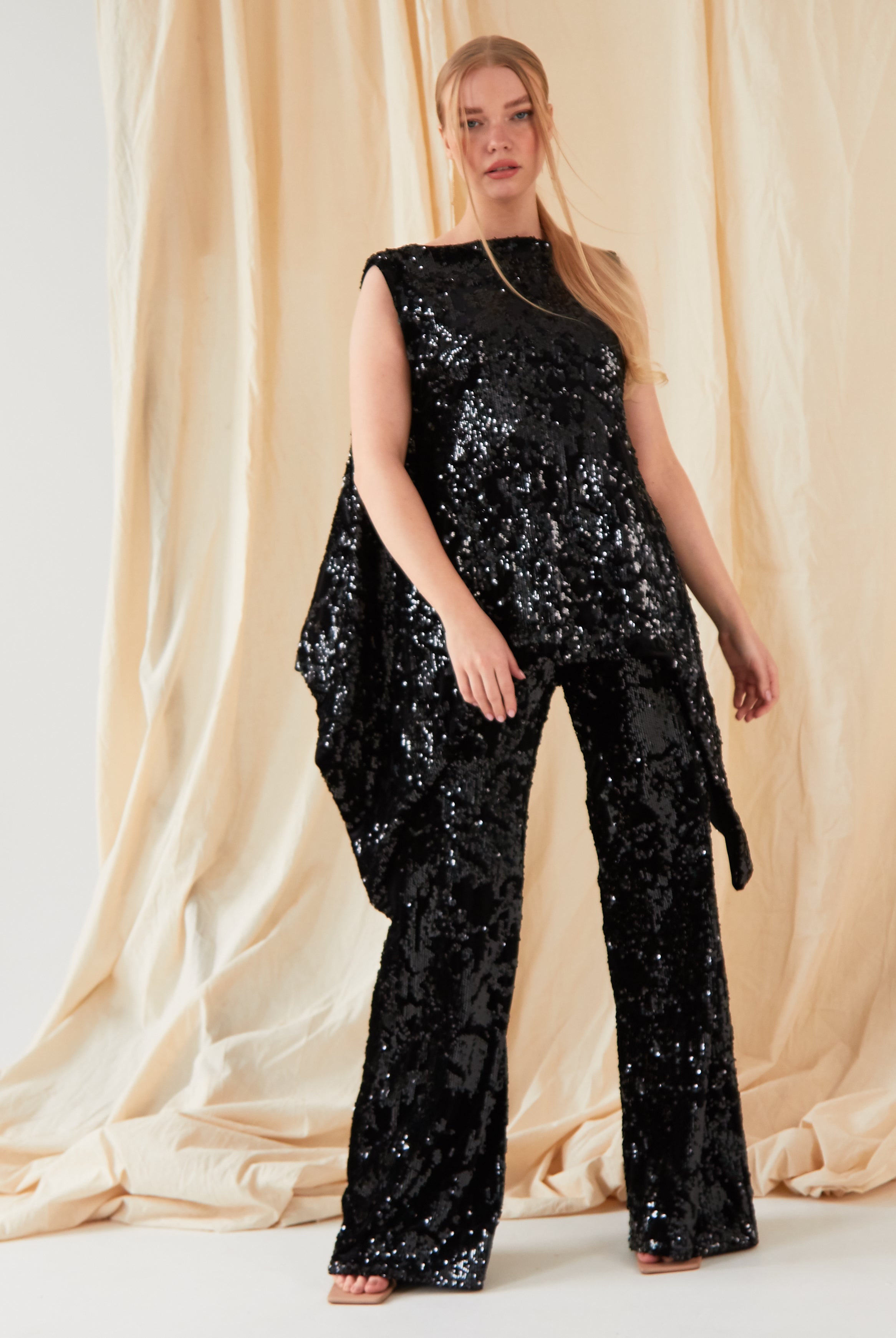 A person with long, straight blonde hair stands before beige drapes, facing the camera in a sleeveless black sequin top and Sarvin’s handmade Sequin Velvet Flared Trousers.