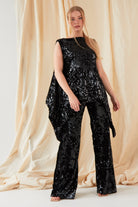 A person with long, straight blonde hair stands before beige drapes, facing the camera in a sleeveless black sequin top and Sarvin’s handmade Sequin Velvet Flared Trousers.