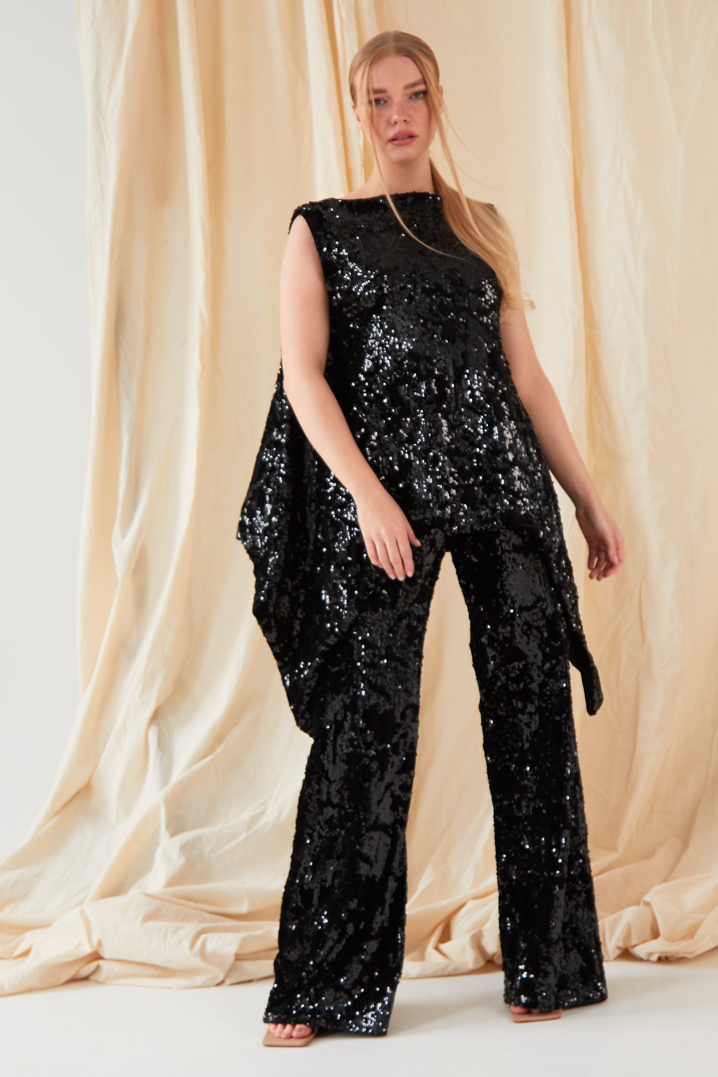 A person with long, straight blonde hair stands before beige drapes, facing the camera in a sleeveless black sequin top and Sarvin’s handmade Sequin Velvet Flared Trousers.