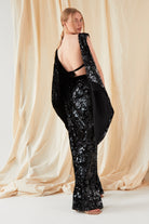 A person stands facing slightly away, wearing Sarvin’s Sequin Velvet Flared Trousers and a black sequined backless top with cape detail against beige draped fabric. This standout outfit is handmade in the UK.