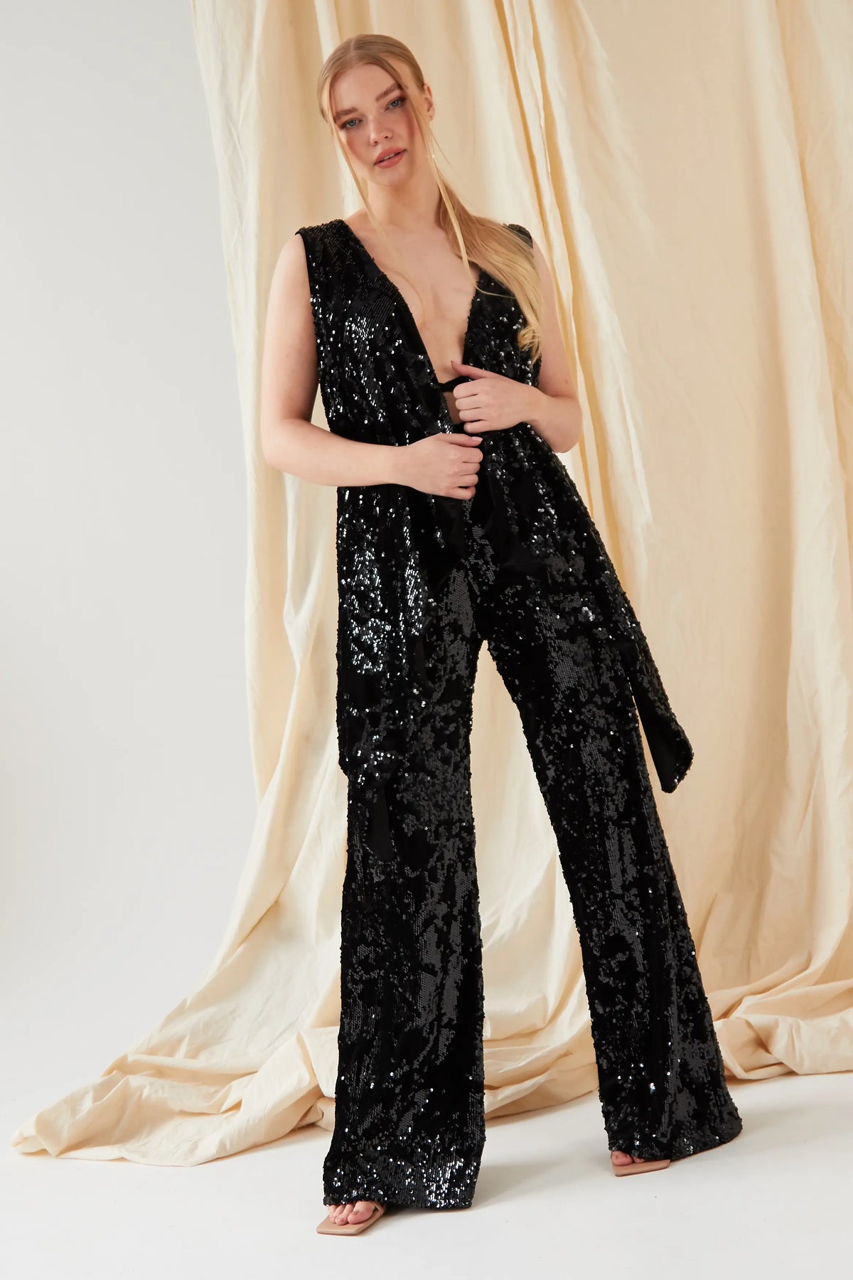 A woman with long blonde hair in a low ponytail stands before beige draped fabric, wearing Sarvin Sequin Velvet Flared Trousers—handmade in the UK—as part of a sleeveless black jumpsuit with a deep V-neck and open-toe shoes.
