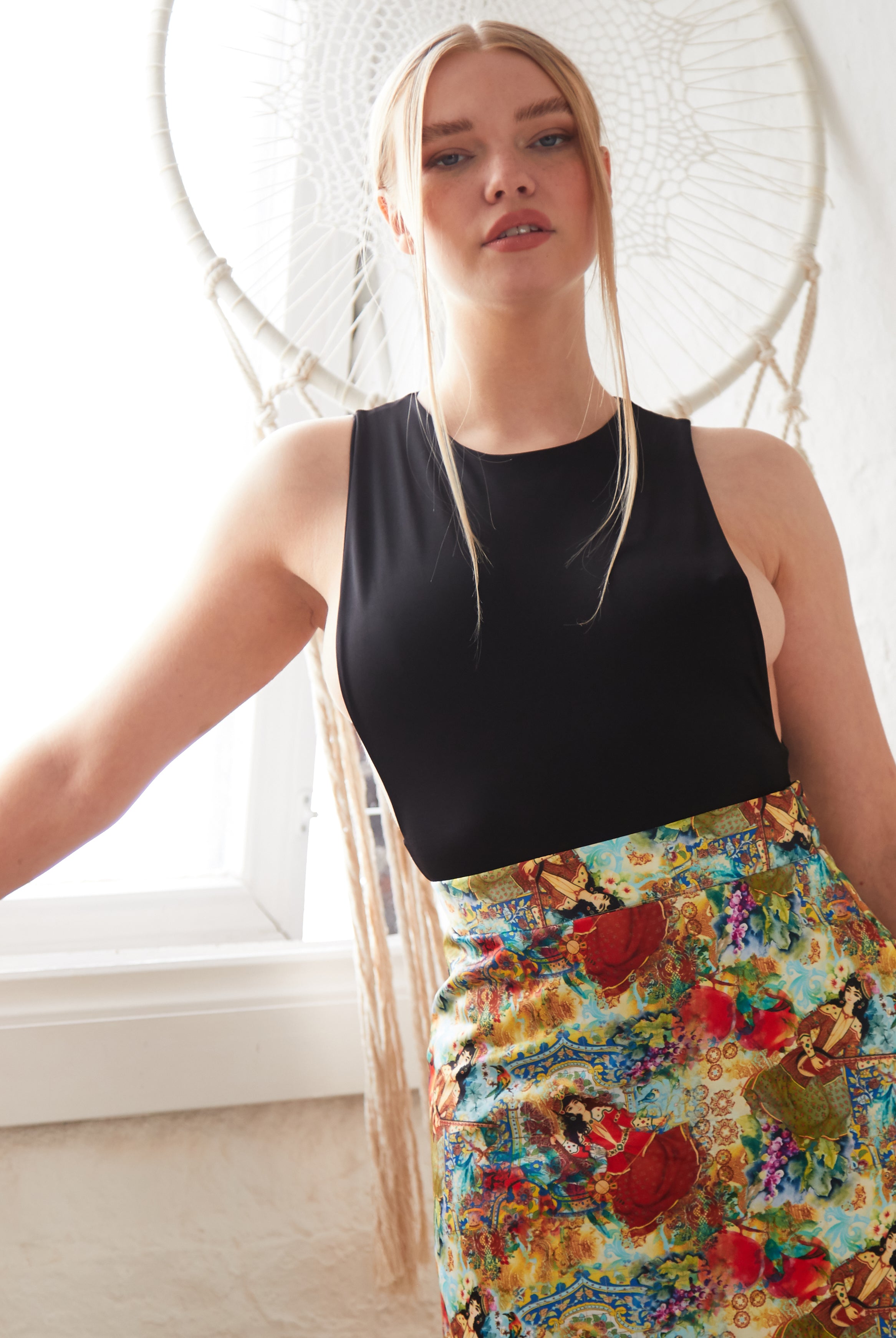 A woman with long blonde hair wears a sleeveless black top and the Sarvin Persian Printed Mini Skirt, posing indoors by a window and a large decorative dreamcatcher.