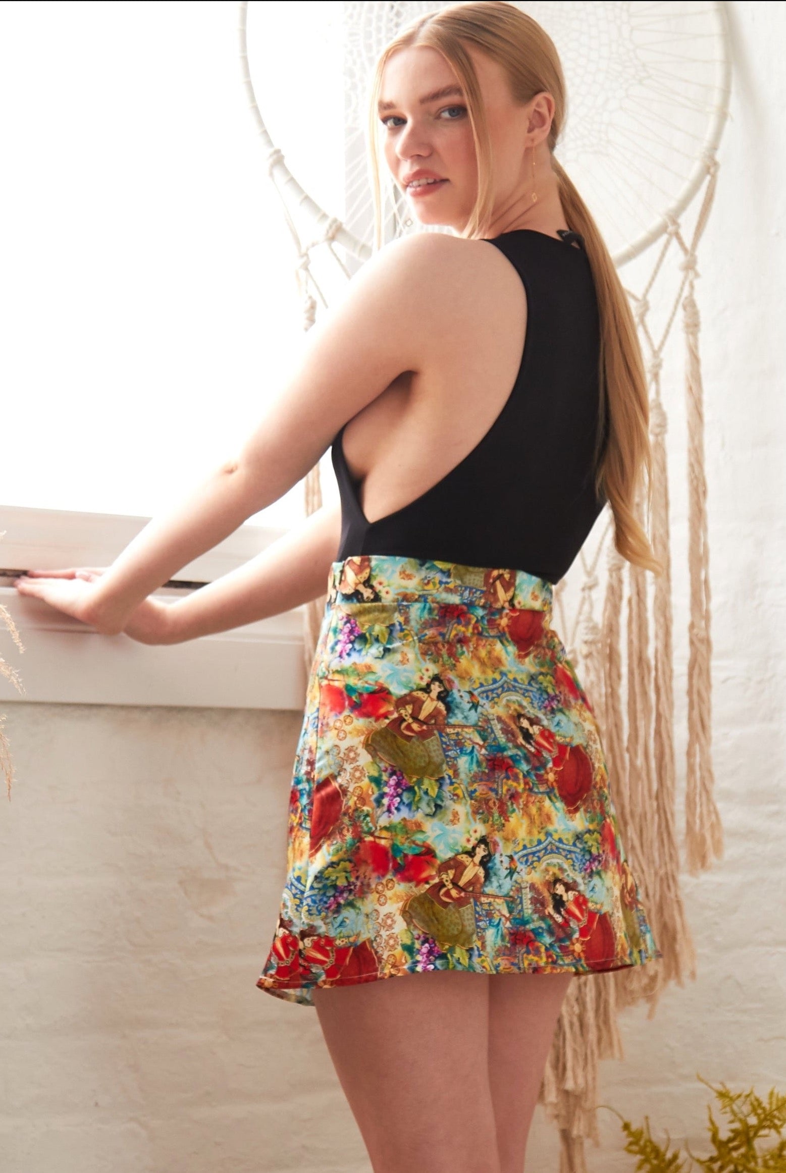 A woman with long blonde hair stands by a window in a sleeveless black top and the Sarvin Persian Printed Mini Skirt, looking over her shoulder at the camera. The background features dried plants and a macramé wall hanging.