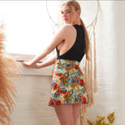A woman with long blonde hair stands by a window in a sleeveless black top and the Sarvin Persian Printed Mini Skirt, looking over her shoulder at the camera. The background features dried plants and a macramé wall hanging.
