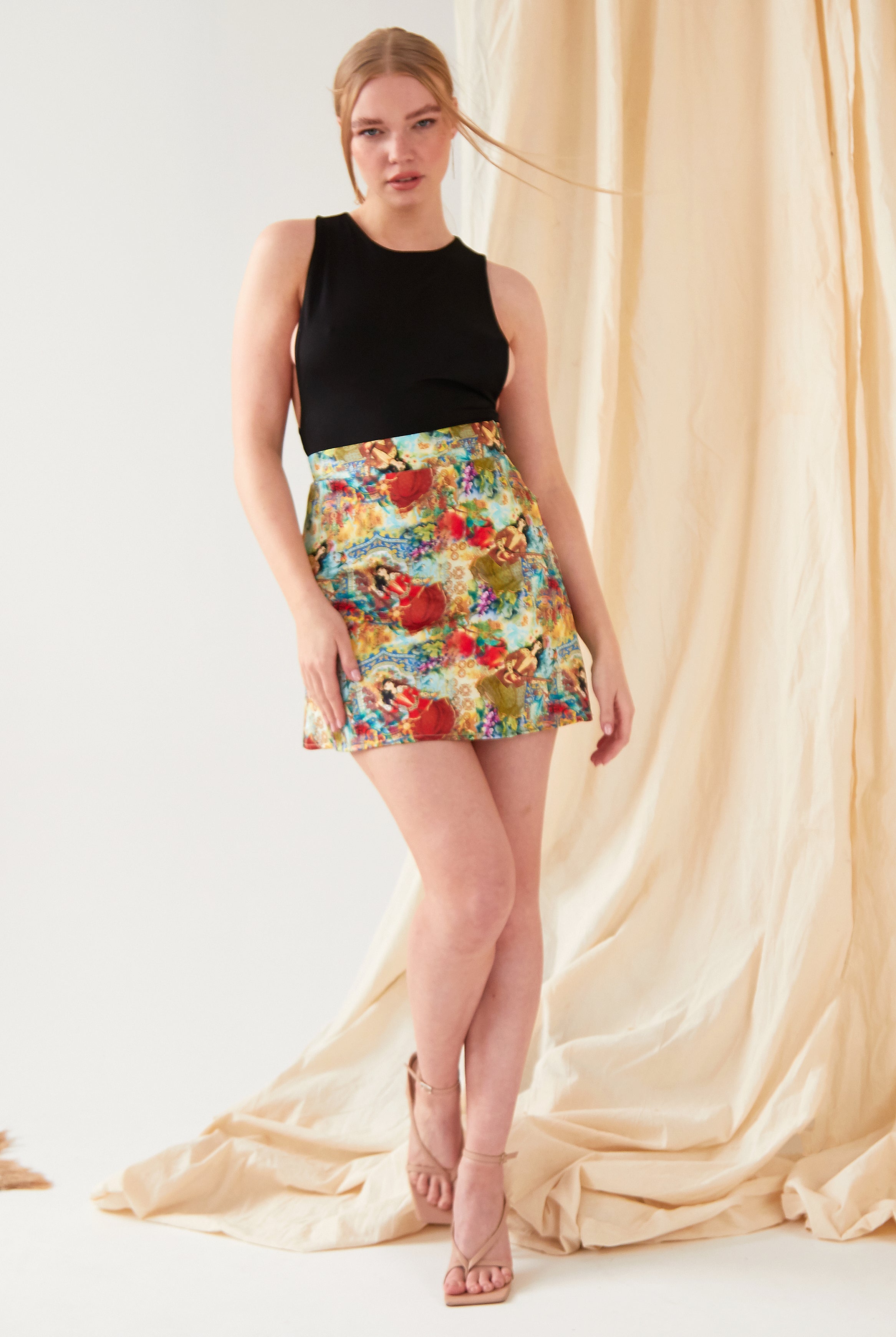 A woman with long blonde hair tied back stands before beige draped fabric, wearing Sarvin's Persian Printed Mini Skirt and a sleeveless black top, styled with nude high-heeled sandals.