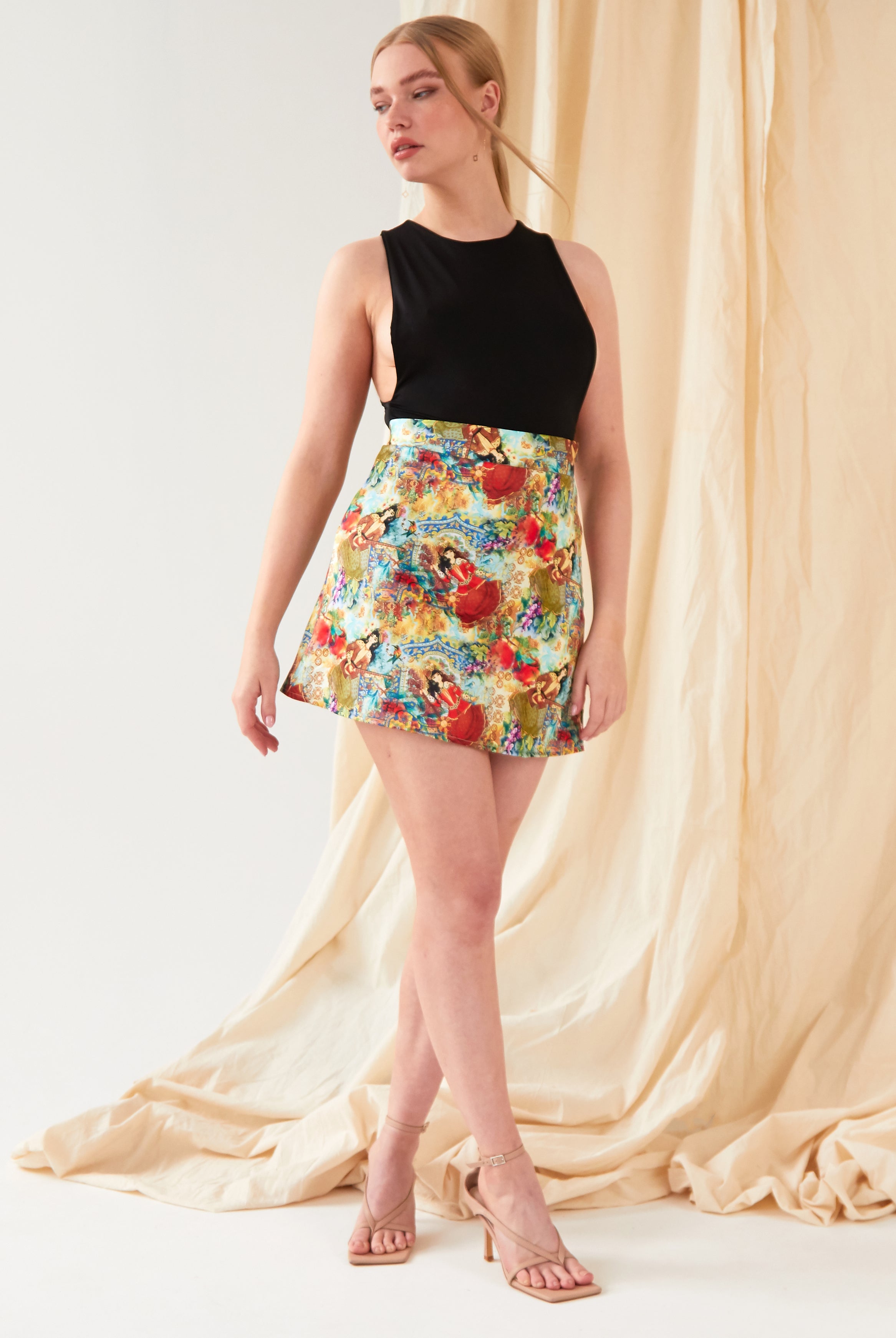 A woman with light hair tied back poses before beige draped fabric, wearing Sarvin’s Persian Printed Mini Skirt and a sleeveless black top, styled with nude heeled sandals.