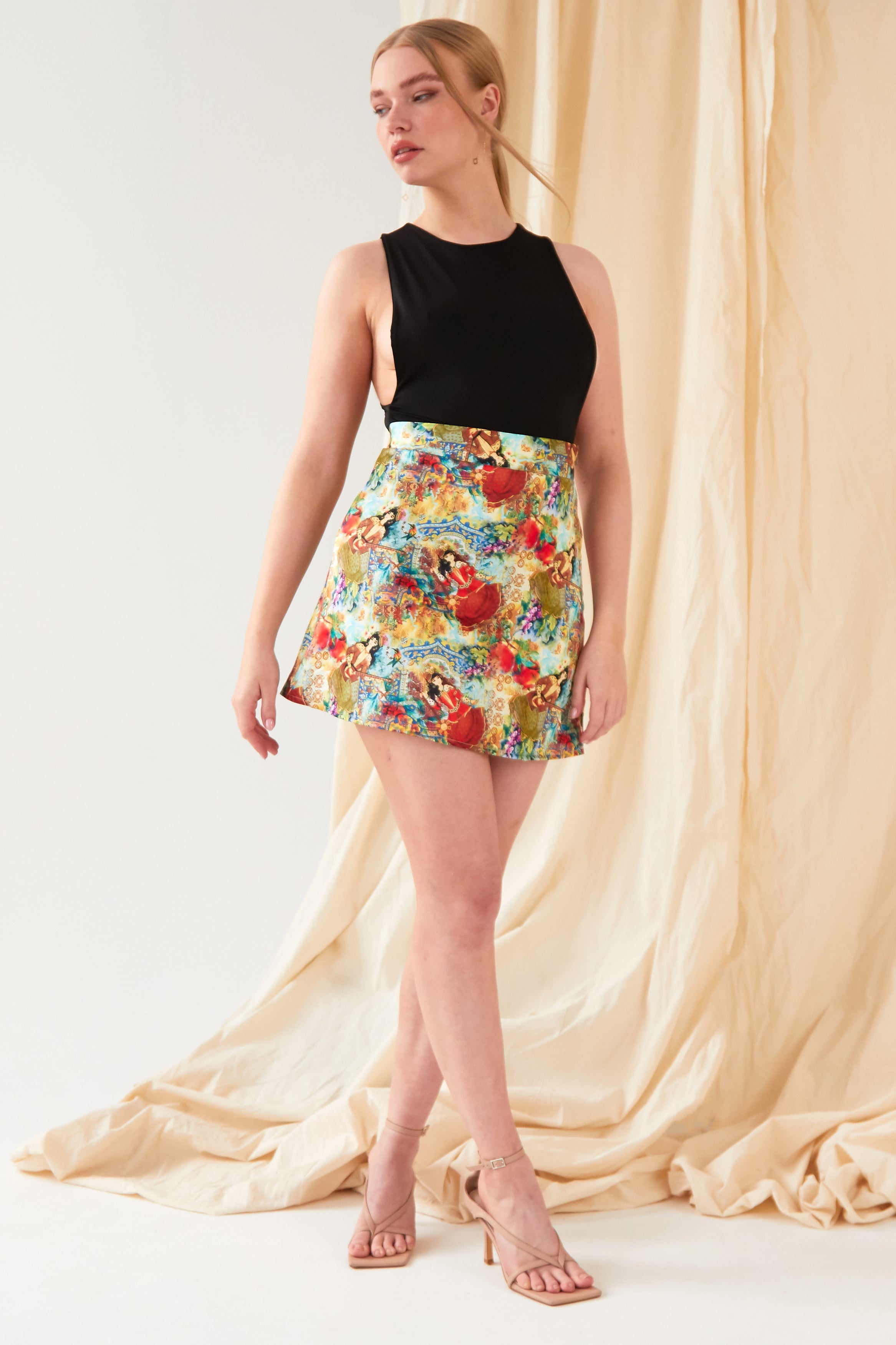 A woman with light hair tied back poses before beige draped fabric, wearing Sarvin’s Persian Printed Mini Skirt and a sleeveless black top, styled with nude heeled sandals.