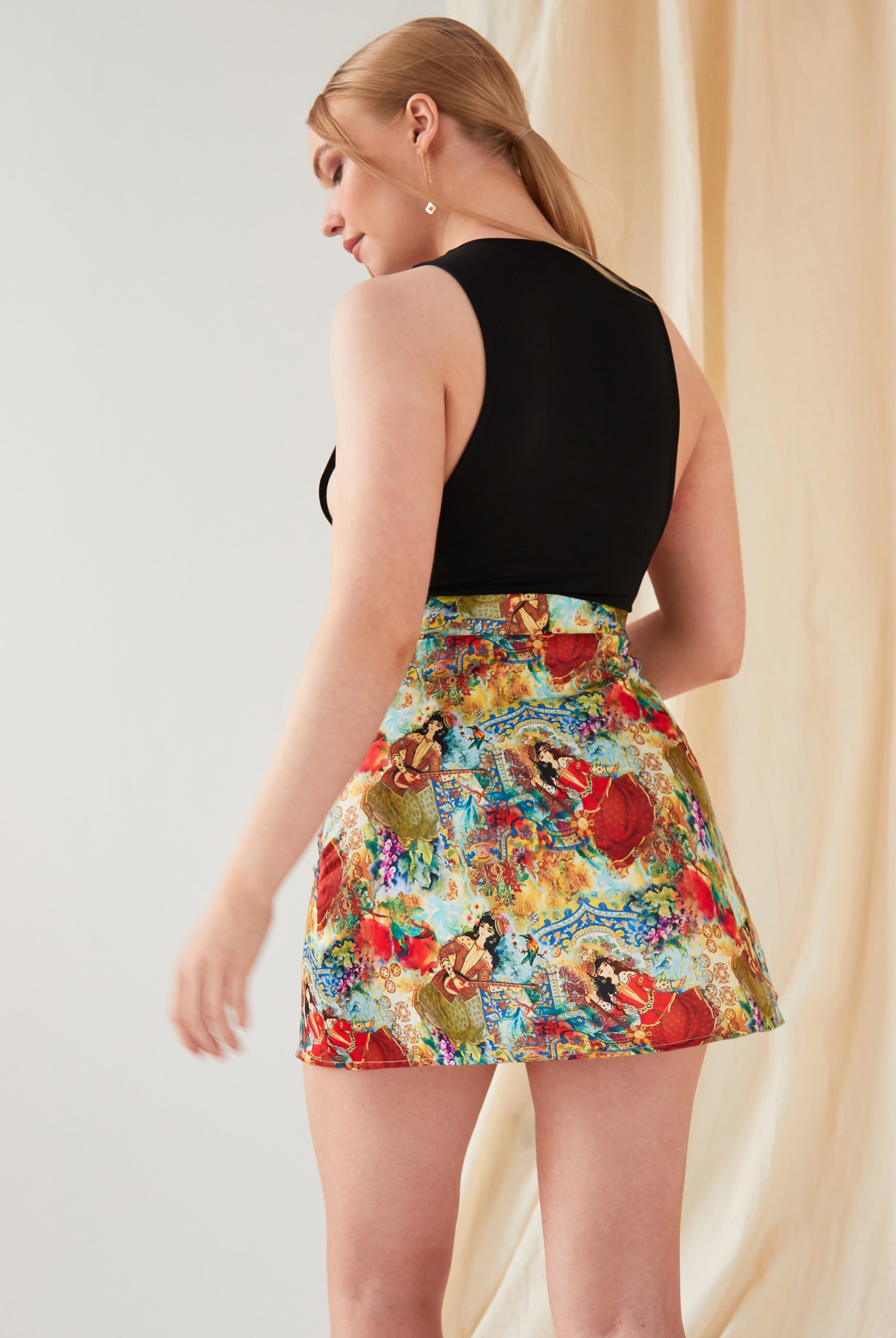 A light-skinned, blonde-haired person faces away, wearing a sleeveless black top and the Sarvin Persian Printed Mini Skirt. A light beige curtain is in the background.