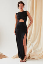 A person poses indoors wearing the Sarvin Black Cut-Out Maxi Dress, featuring a high slit, paired with strappy sandals, against beige sparkly draped fabric.