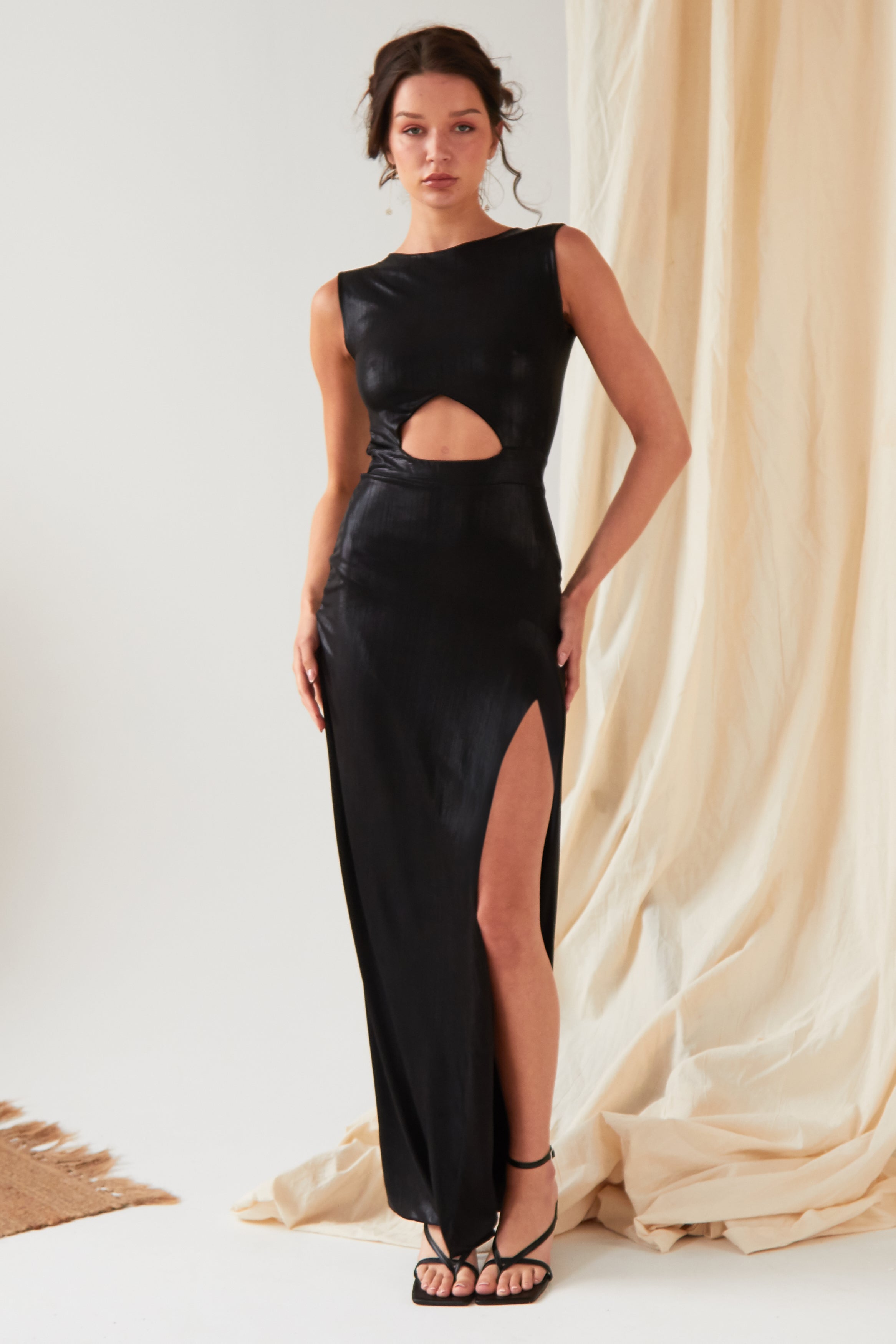 A person poses indoors wearing the Sarvin Black Cut-Out Maxi Dress, featuring a high slit, paired with strappy sandals, against beige sparkly draped fabric.