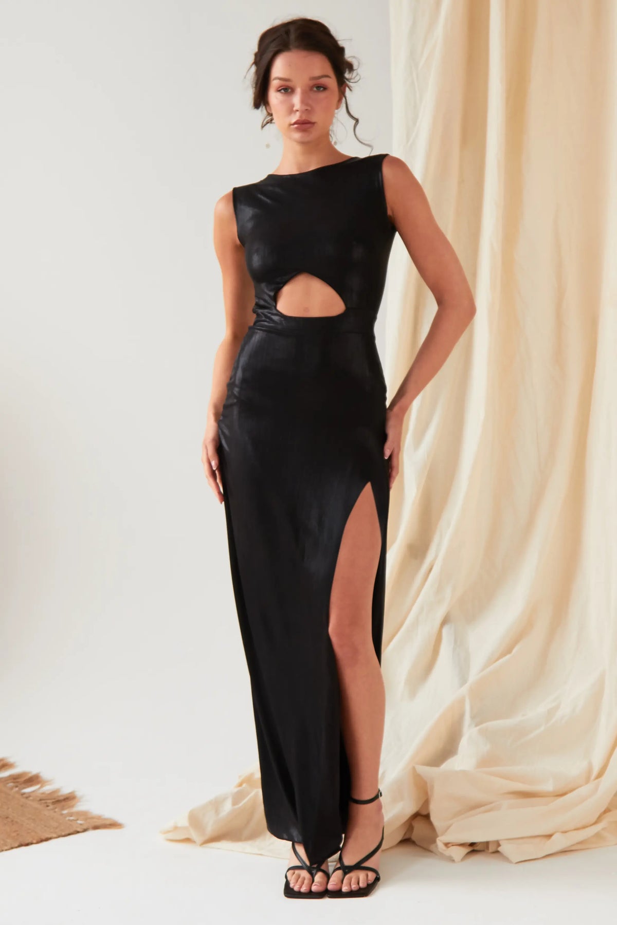 A person poses indoors wearing the Sarvin Black Cut-Out Maxi Dress, featuring a high slit, paired with strappy sandals, against beige sparkly draped fabric.