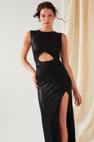 A person is wearing the Sarvin Black Cut-Out Maxi Dress, featuring a front waist cutout and high slit, while standing in front of light-colored draped sparkly fabric.